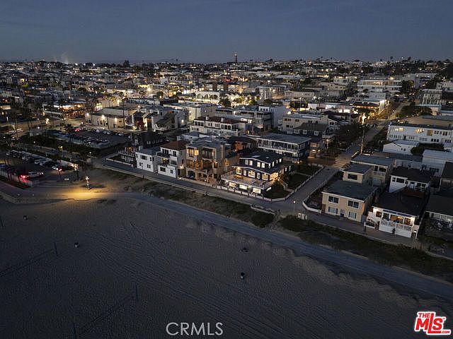 This high-angle aerial view captures a coastal neighborhood at twilight, showcasing a dense collection of homes situated directly along a sandy beach. The perspective highlights the proximity of the properties to the ocean, with street lights and house lights creating a warm, inviting glow against the darkening sky. The scene emphasizes the prime location and the layout of the beachfront community.