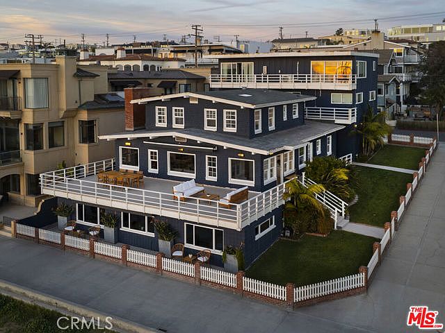This elevated aerial perspective captures a multi-level, dark-blue coastal residence featuring a prominent wrap-around deck furnished with outdoor seating. The property includes a white picket fence enclosing a manicured lawn and is situated in a dense neighborhood near the coast. The architectural style emphasizes outdoor living spaces and tiered levels, providing a clear view of the home's exterior layout and its relationship to the surrounding streetscape.