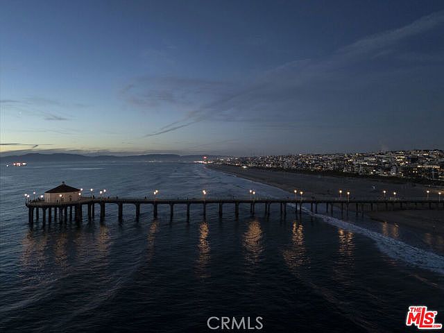 This aerial view captures a long pier extending into the ocean at twilight, with the coastline and city lights stretching into the distance. The scene features a prominent pier-end building illuminated by warm lights, creating a serene and picturesque atmosphere. The perspective is wide and cinematic, showcasing the expansive horizon and the calm, dark waters of the Pacific.
