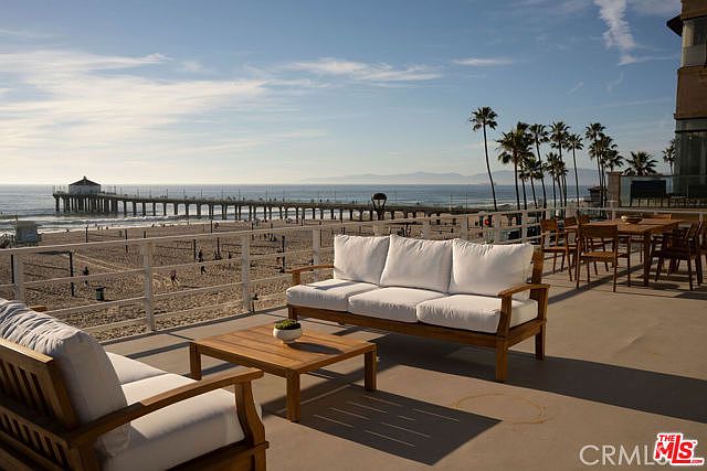 This expansive outdoor deck offers a stunning, elevated view of the ocean and a prominent pier. The space is furnished with comfortable light-wood seating featuring white cushions, arranged around a matching coffee table, creating an inviting lounge area. Beyond the railing, the sandy beach and the vast horizon provide a serene and picturesque backdrop for this coastal property.
