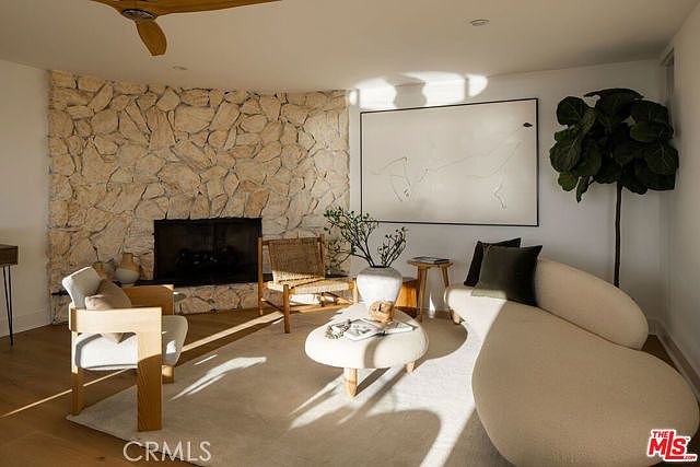 This bright living room features a prominent stone-clad chimney breast that serves as a focal point, complemented by light wood flooring and a neutral color palette. The space is furnished with a curved, cream-colored sofa, a modern wooden armchair, and a low, rounded coffee table, creating a soft and organic aesthetic. A large, minimalist piece of art hangs on the wall, while a tall potted plant adds a touch of greenery to the corner.