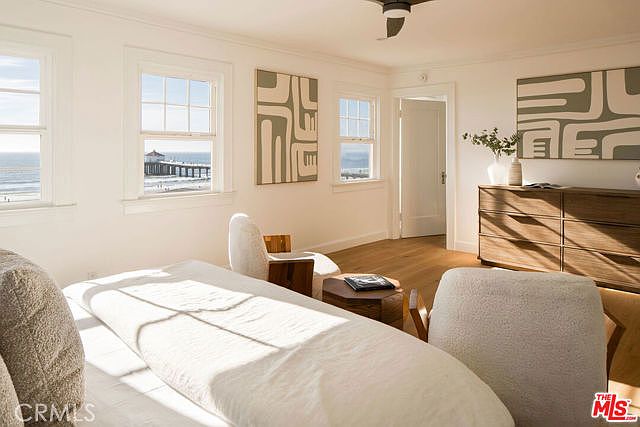 This bright and airy bedroom features light wood flooring and a neutral color palette, creating a serene coastal atmosphere. The room is furnished with two textured armchairs, a wooden dresser, and a bed, while large windows provide a direct, picturesque view of the ocean and a pier. Abstract wall art adds a modern touch to the space, which is illuminated by natural light streaming in from the windows.