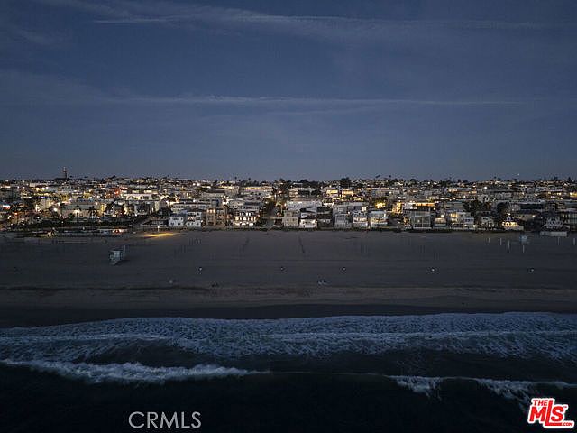 This aerial view captures a coastal neighborhood at twilight, showcasing a long stretch of sandy beach meeting the ocean waves. The scene features a dense collection of residential properties illuminated by warm interior and exterior lights, creating a vibrant contrast against the darkening sky. The perspective provides a wide, cinematic look at the property's prime location relative to the shoreline.