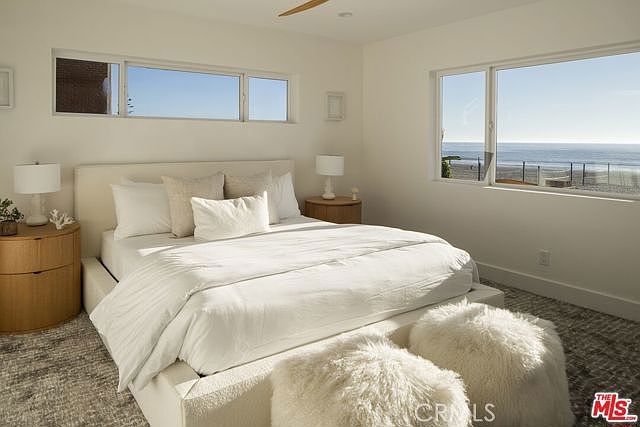 This bright and airy primary bedroom features a neutral color palette and a large bed centered in the room, flanked by two round wooden nightstands with matching lamps. A pair of plush, white faux-fur ottomans sits at the foot of the bed, adding texture to the space. Large windows provide an expansive, unobstructed view of the ocean and beach, creating a serene and luxurious coastal atmosphere.