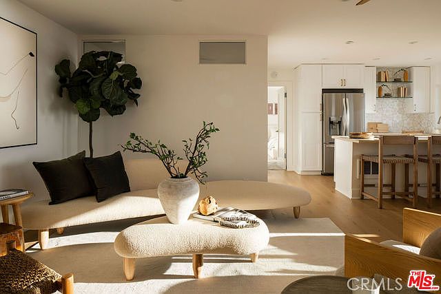 This bright and airy living room features a minimalist, organic aesthetic with a curved, cream-colored sofa and a matching sculptural coffee table. The space flows seamlessly into an open-concept kitchen equipped with light wood cabinetry and a central island with bar seating. Natural light floods the room, highlighting the light wood flooring and creating a warm, inviting atmosphere.
