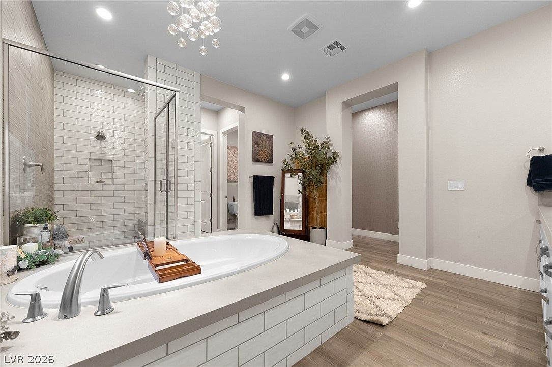 This primary bathroom features a spacious soaking tub set into a tiled platform, adjacent to a large walk-in shower with light-colored subway tile walls. The room is accented by a crystal chandelier, a tall decorative mirror, and light wood-toned flooring that extends throughout the space. The neutral color palette and clean lines create a bright, spa-like atmosphere.