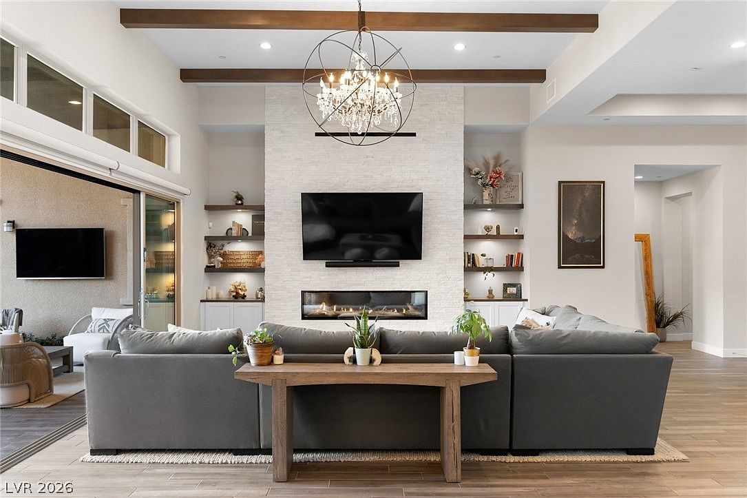 This spacious living room features a prominent stone-clad chimney breast centered between built-in shelving units, housing a linear fireplace and a wall-mounted television. A large, dark gray sectional sofa faces the fireplace, accented by a light wood console table and several potted plants. The room is illuminated by a decorative chandelier suspended from a ceiling with exposed wood beams, while large sliding glass doors on the left provide a view of the outdoor space.