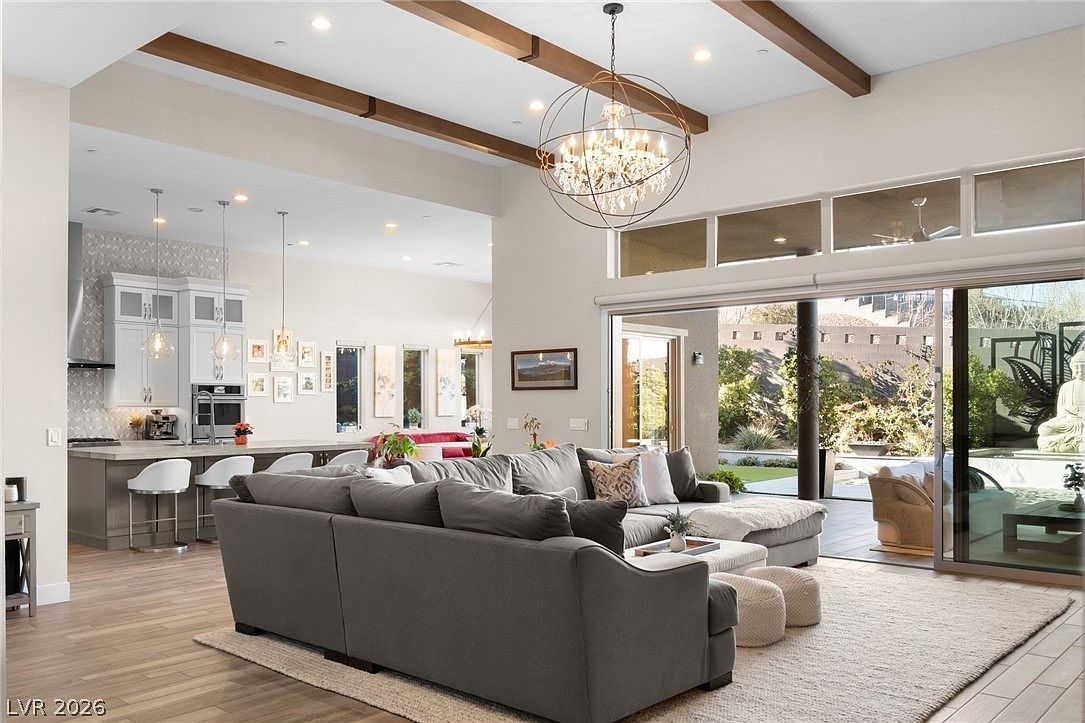 This open-concept living area features a large, plush sectional sofa centered on a textured area rug, creating a comfortable space for relaxation. The room is highlighted by exposed wooden ceiling beams and a striking, spherical chandelier, while large sliding glass doors provide a seamless transition to the outdoor patio. In the background, a bright, modern kitchen with a breakfast bar and pendant lighting completes the airy, sophisticated layout.