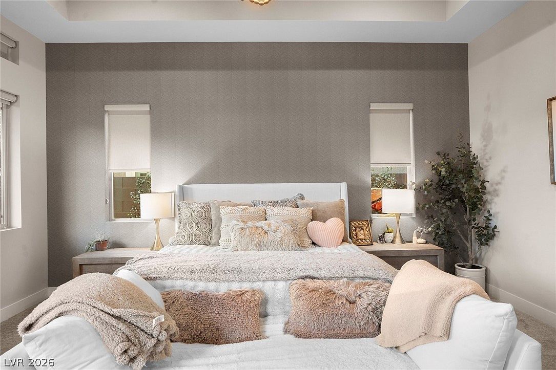 This primary bedroom features a neutral color palette with a textured, patterned accent wall behind a white upholstered bed frame. The room is styled with plush, layered bedding, including decorative pillows and a heart-shaped accent cushion, flanked by two matching nightstands with elegant table lamps. A potted plant adds a touch of greenery to the corner, while two windows with roller shades allow natural light to brighten the space.