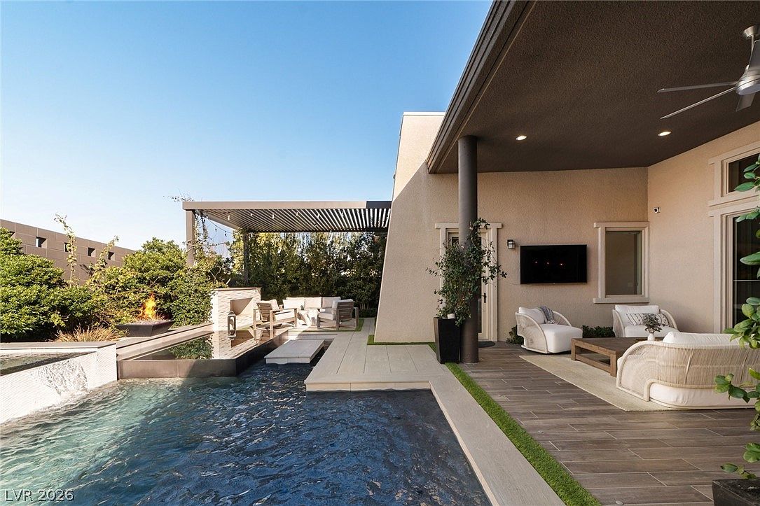 This luxurious outdoor living space features a covered patio area with comfortable lounge seating and a wall-mounted television, seamlessly transitioning to a poolside deck. The area includes a modern fire feature and a sleek, dark-tiled swimming pool, all set against a backdrop of lush greenery and a contemporary pergola. The design emphasizes a sophisticated indoor-outdoor lifestyle, perfect for entertaining in a private, resort-style setting.