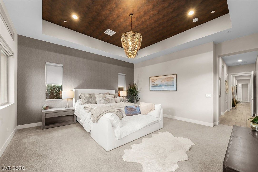 This spacious primary bedroom features a dramatic tray ceiling with dark wood-toned paneling and a decorative beaded chandelier. The room is anchored by a large upholstered bed with a plush bench at the foot, set against a textured accent wall. A soft area rug sits atop the carpeted floor, and the neutral color palette creates a serene and sophisticated atmosphere.