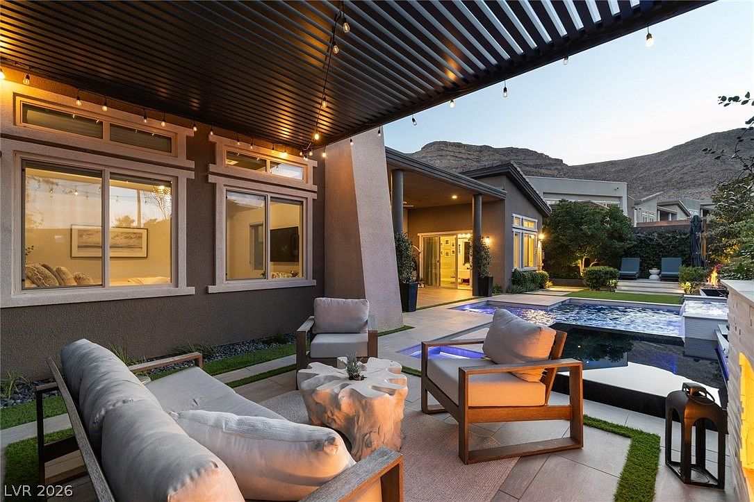 This inviting outdoor living space features a covered patio area furnished with a comfortable sofa, armchairs, and a unique organic-shaped wood coffee table. The space is illuminated by string lights hanging from a slatted pergola, creating a warm ambiance that overlooks a modern swimming pool and spa. The backdrop includes a desert landscape with mountain views, emphasizing a luxurious and relaxing lifestyle.
