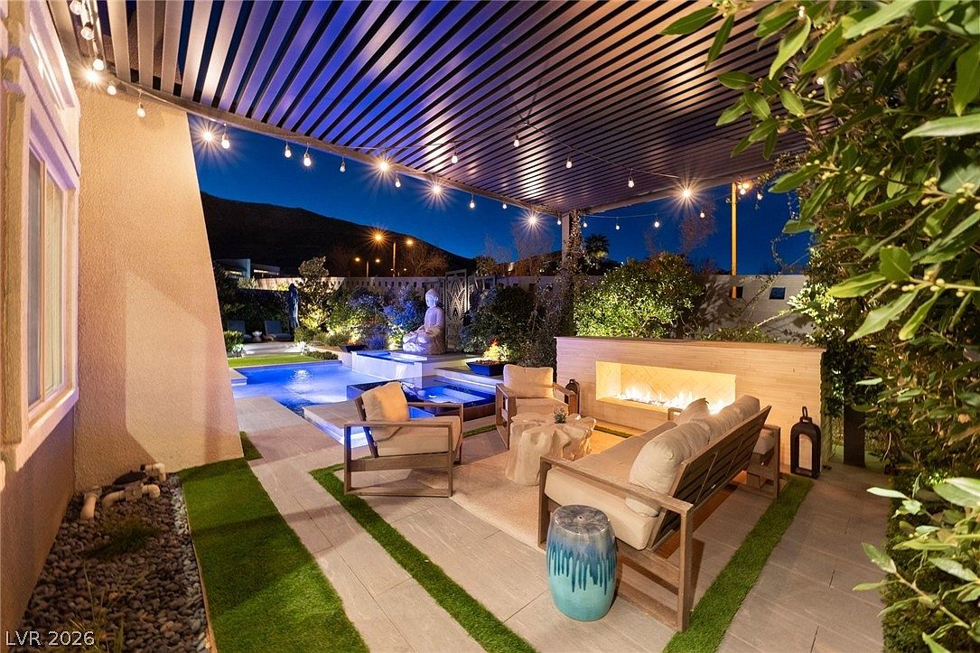 This inviting outdoor living space features a covered patio area with a built-in fireplace and comfortable lounge seating, perfect for evening relaxation. The space transitions seamlessly to a swimming pool and spa, accented by a decorative Buddha statue and lush landscaping. String lights suspended from the overhead structure create a warm, ambient atmosphere against the twilight sky.