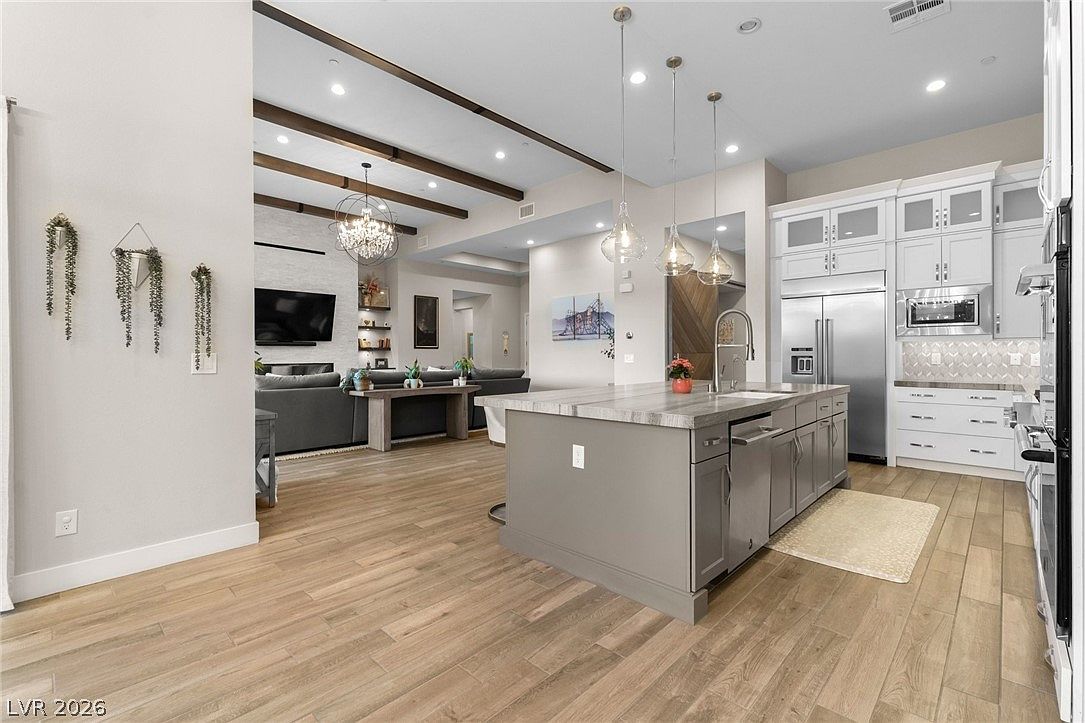 This open-concept kitchen features a large central island with a grey base and a light-toned countertop, serving as a focal point for the space. The kitchen is equipped with white cabinetry, stainless steel appliances, and a tiled backsplash, while the adjacent living area is visible through an open transition defined by dark wood ceiling beams. The perspective captures the seamless flow between the culinary workspace and the comfortable living room, highlighting a bright, modern, and inviting home layout.