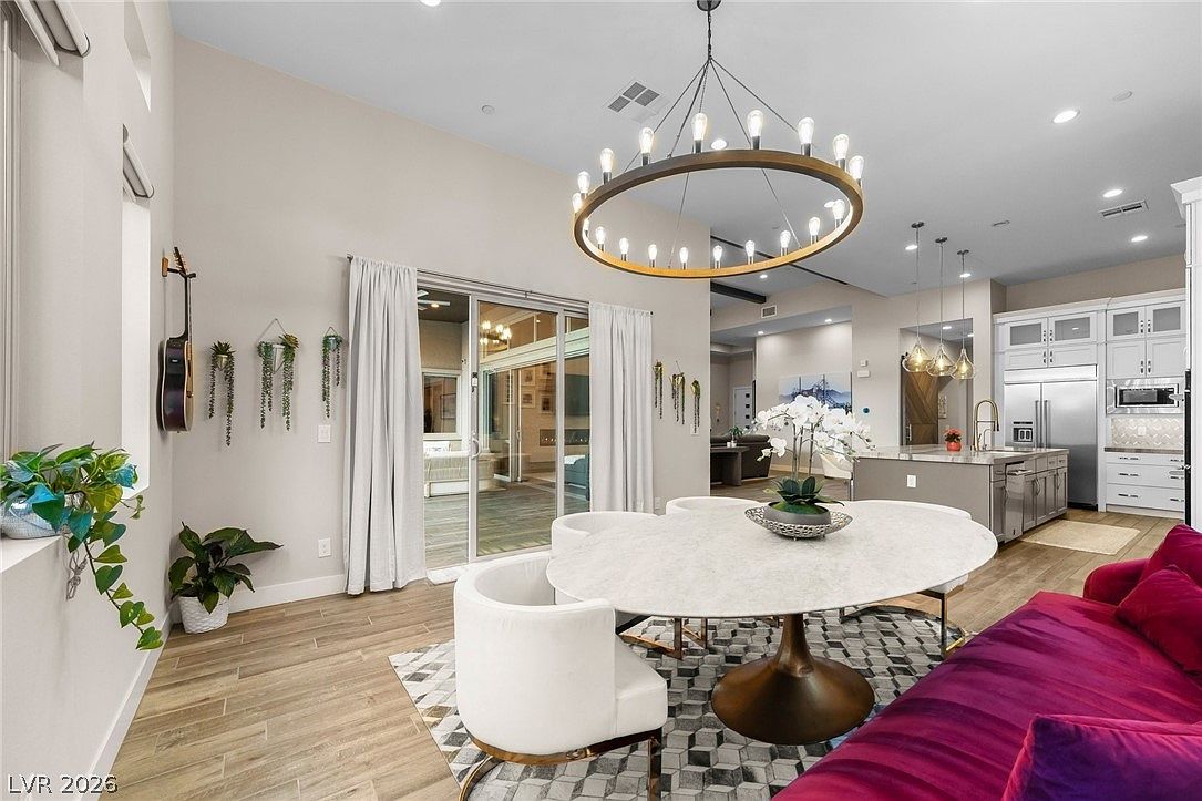 This open-concept dining area features a modern round table with white upholstered chairs, positioned beneath a large, circular chandelier with multiple exposed bulbs. The space flows seamlessly into a bright kitchen with light-toned cabinetry and a central island, while a vibrant magenta sofa adds a bold pop of color to the foreground. Large sliding glass doors provide a clear view of an outdoor patio, enhancing the airy and connected feel of the home.
