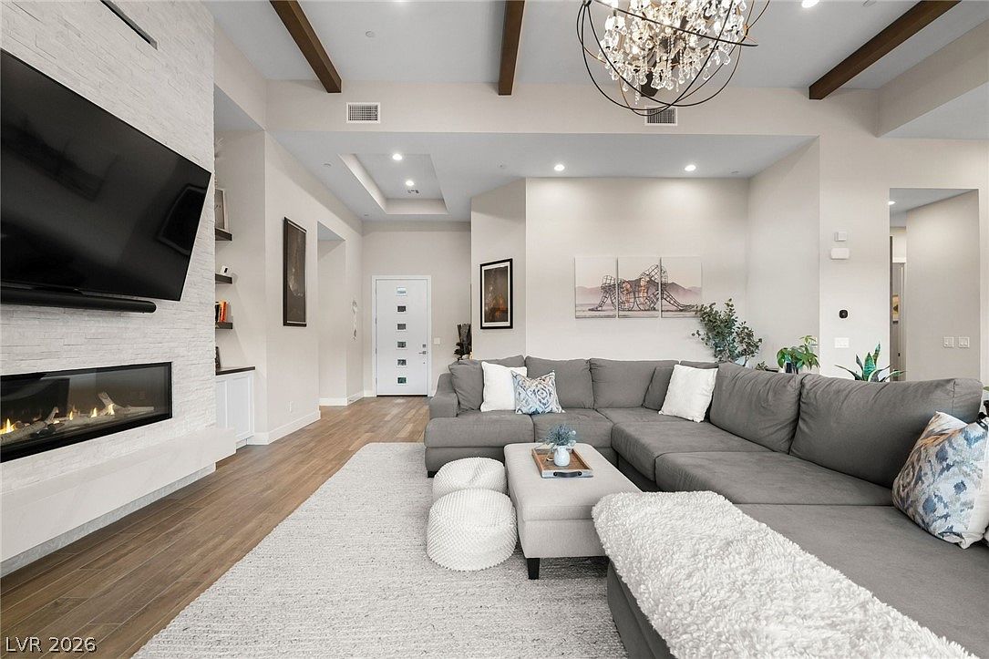 This spacious living room features a large gray sectional sofa arranged around a central ottoman and two round poufs, all resting on a textured area rug. A prominent stone-clad chimney breast houses a wide, linear fireplace beneath a wall-mounted television, while exposed wood beams accent the high ceiling. The room is illuminated by a decorative crystal chandelier and recessed lighting, creating a bright and inviting atmosphere.