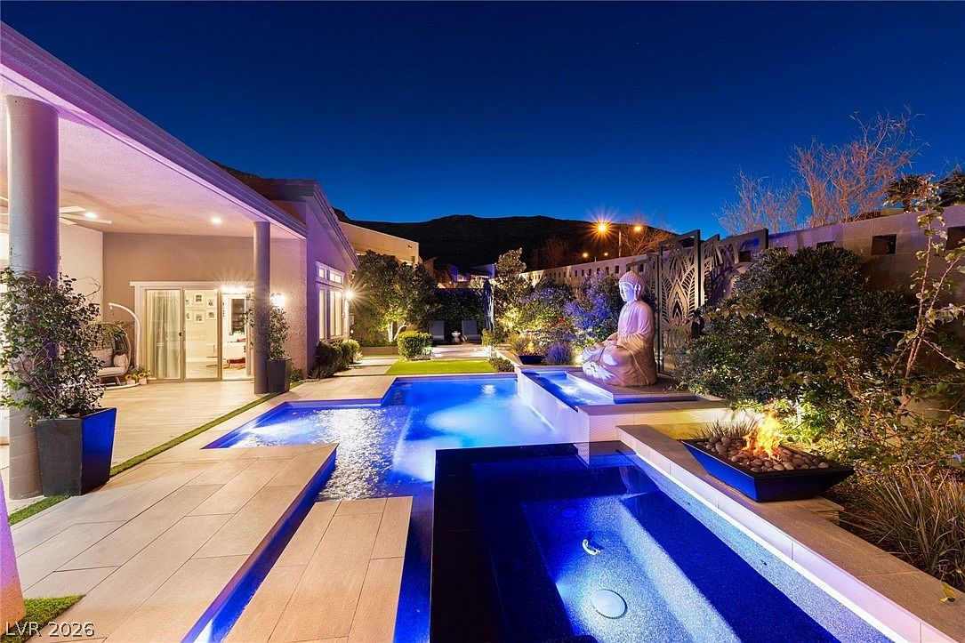 This stunning backyard oasis features a modern swimming pool and integrated spa illuminated by vibrant blue underwater lighting. A serene Buddha statue sits on a raised platform beside a linear fire feature, creating a tranquil and luxurious atmosphere. The scene is captured at twilight, highlighting the seamless transition between the covered patio area and the landscaped outdoor living space.