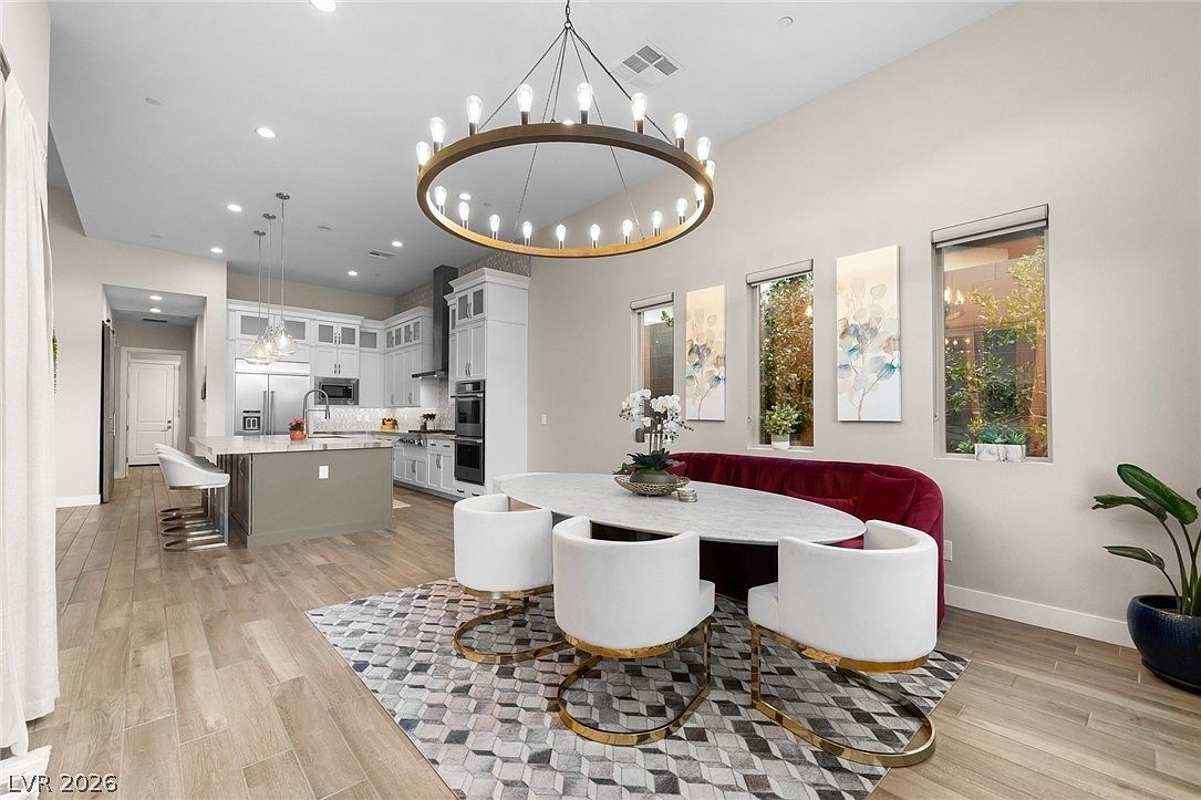 This open-concept dining area flows seamlessly into a modern kitchen, featuring a large oval table paired with white upholstered chairs and a plush, deep red velvet bench. A striking circular chandelier serves as a focal point above the table, while a geometric patterned rug defines the space against the light-toned wood flooring. The adjacent kitchen is equipped with white cabinetry, a central island with bar seating, and stainless steel appliances, creating a cohesive and sophisticated living environment.