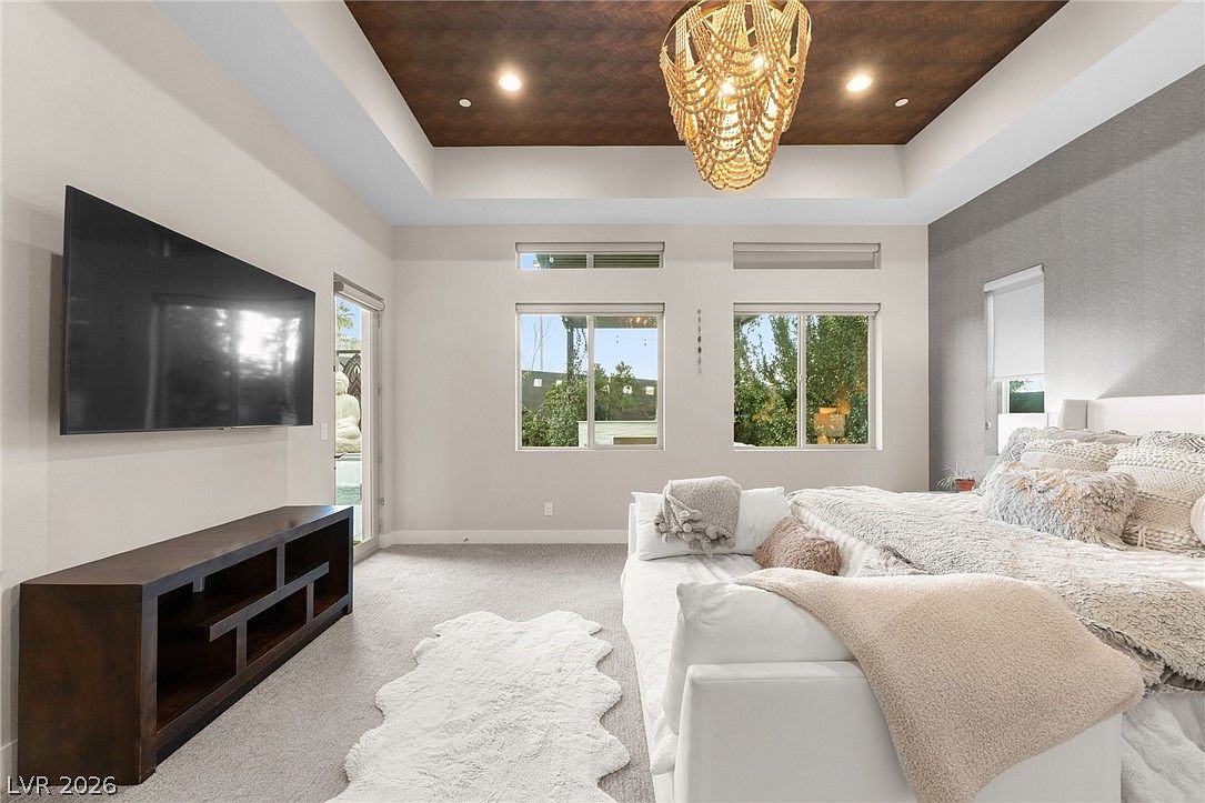 This spacious primary bedroom features a tray ceiling with a dark wood finish and a prominent beaded chandelier. The room is anchored by a large white bed with textured bedding, complemented by a plush white rug and a dark wood media console beneath a wall-mounted television. Large windows allow natural light to fill the space, creating an airy and sophisticated atmosphere.