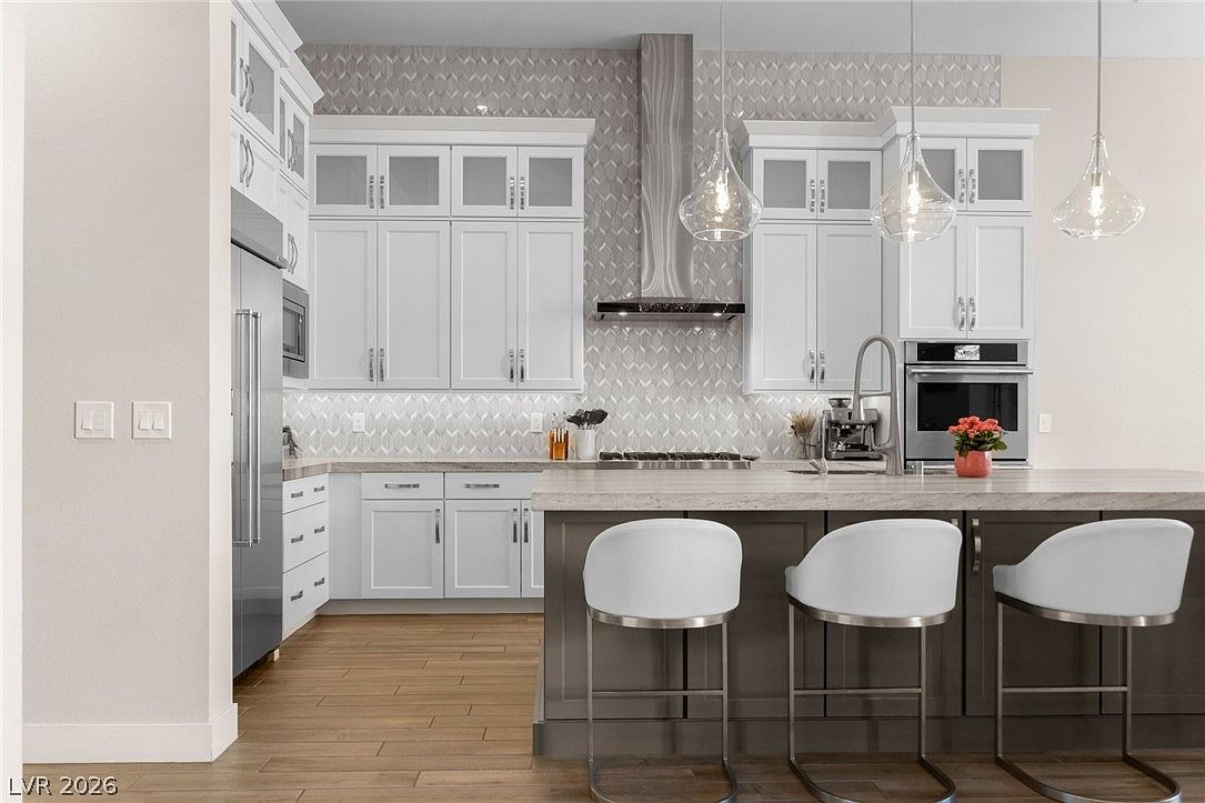 This modern kitchen features crisp white cabinetry contrasted by a dark gray central island with bar seating. A patterned tile backsplash extends behind the stainless steel range hood, while light-toned wood flooring runs throughout the space. The room is illuminated by three glass pendant lights hanging over the island, creating a bright and inviting atmosphere.
