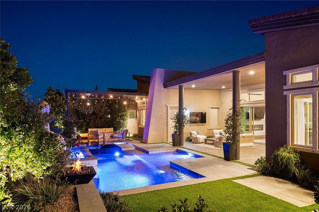 This evening shot captures a luxurious backyard oasis featuring a modern swimming pool with integrated lighting and a fire feature. A covered patio area provides an inviting outdoor living space, complete with comfortable seating and a wall-mounted television. The scene is framed by manicured landscaping and string lights, creating a sophisticated and tranquil atmosphere for evening relaxation.