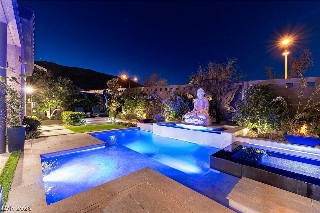 This nighttime exterior shot showcases a luxurious backyard oasis featuring a modern swimming pool with vibrant blue LED lighting and an integrated spa. A prominent Buddha statue serves as a focal point on a raised stone platform, complemented by lush landscaping, accent lighting, and a fire feature. The scene is captured from a low-angle perspective, emphasizing the serene and high-end resort-style atmosphere of the outdoor living space.