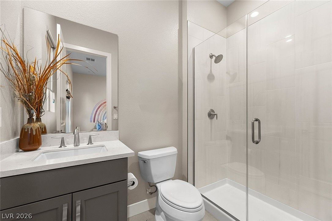 This modern bathroom features a dark gray vanity with a white countertop and a rectangular undermount sink. To the right, a glass-enclosed walk-in shower with light-colored wall tiles and a chrome shower head provides a clean, contemporary aesthetic. A white toilet is positioned between the vanity and the shower, and a large mirror above the sink reflects a decorative rainbow wall art piece.