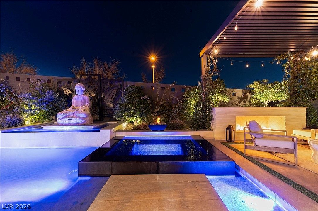 This luxurious outdoor space features a modern, illuminated swimming pool and a central square spa, creating a serene nighttime atmosphere. A prominent Buddha statue sits on a raised platform near the water, complemented by a cozy seating area with an outdoor fireplace and a pergola overhead. The scene is elegantly lit, highlighting the clean lines and sophisticated landscaping, perfect for evening relaxation and entertaining.