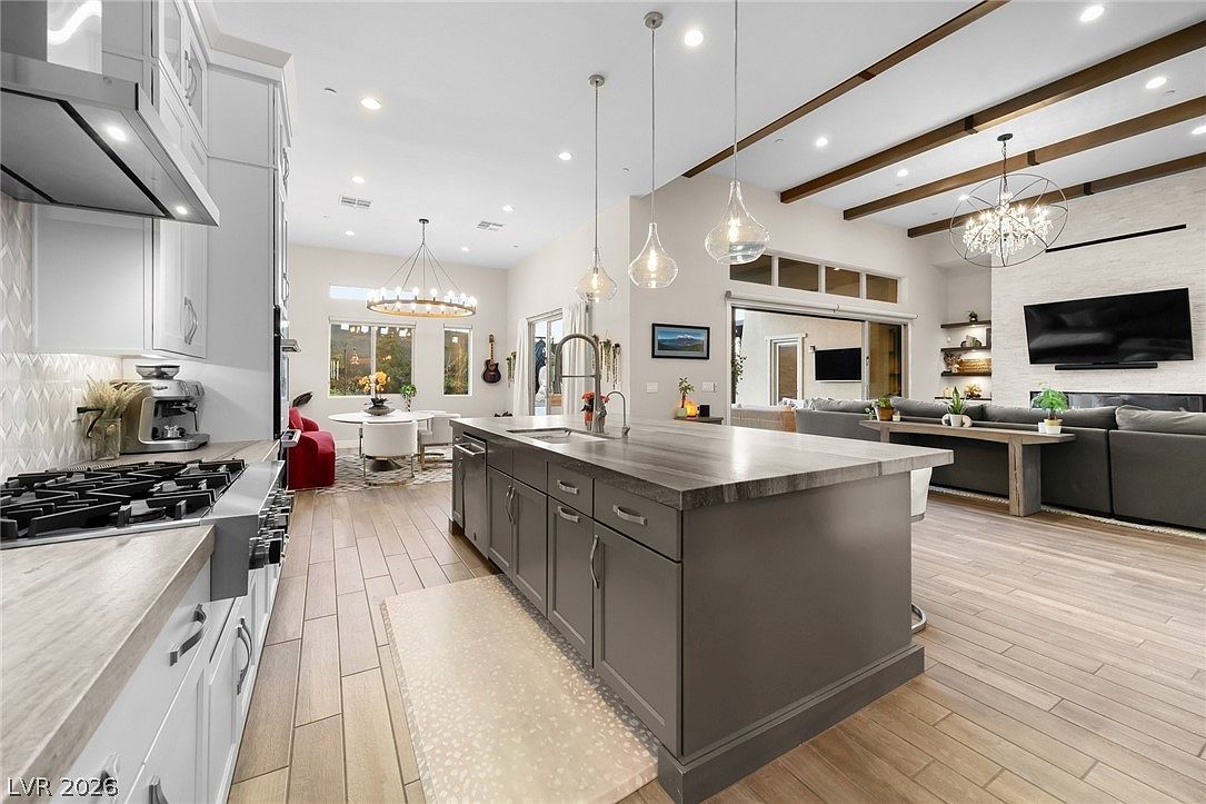 This open-concept kitchen features a large central island with a dark finish and a light-toned countertop, serving as the focal point of the space. The kitchen is equipped with stainless steel appliances, including a gas range and a built-in coffee machine, complemented by white cabinetry and pendant lighting. The perspective looks from the kitchen across the island toward an adjacent dining area and a living room with a stone-clad chimney breast, creating a cohesive and airy living environment.