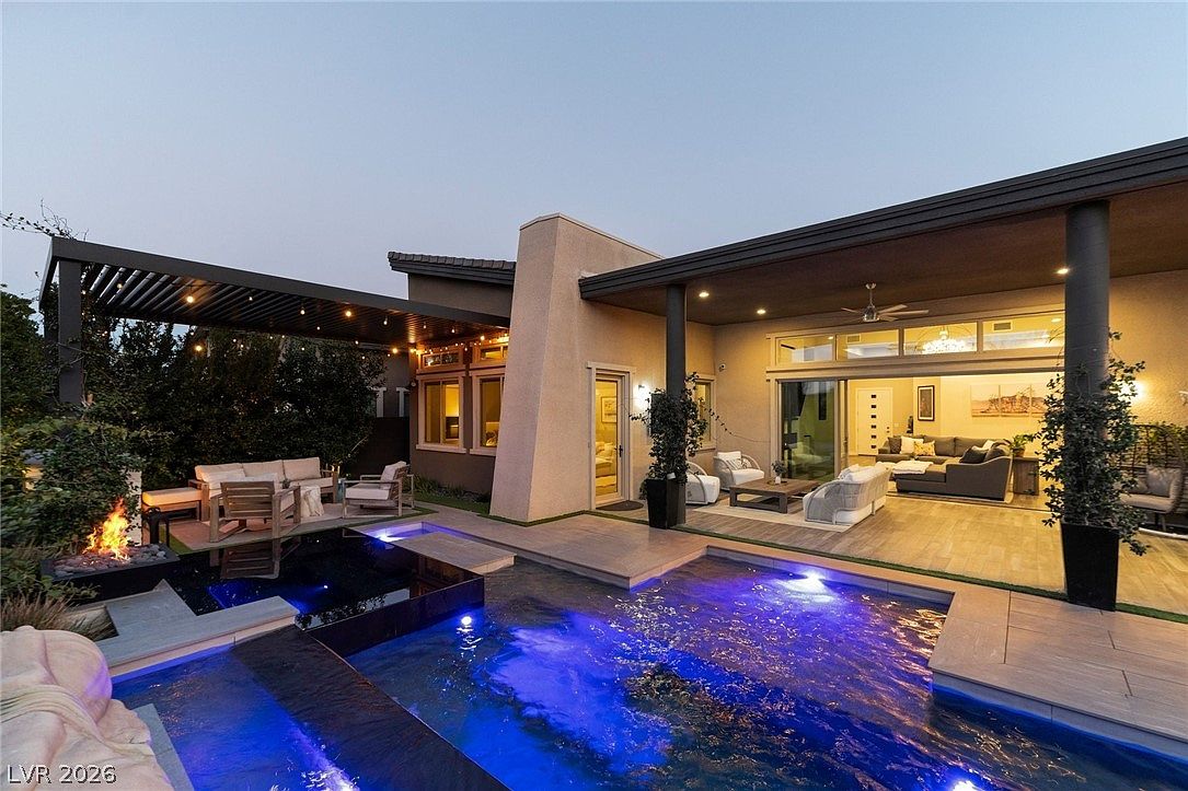 This luxurious backyard features a modern swimming pool with vibrant blue LED lighting and a built-in fire pit area. A spacious covered patio with comfortable outdoor seating seamlessly connects to the interior living space through large sliding glass doors. The architectural design emphasizes indoor-outdoor living, creating a sophisticated and inviting atmosphere for evening relaxation.