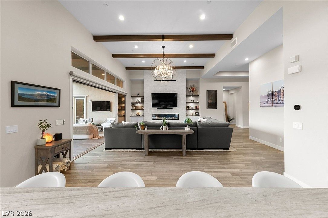 This spacious, open-concept living room features a high ceiling with exposed wooden beams and a central stone-clad chimney breast housing a television and a linear fireplace. A large, plush gray sectional sofa faces the fireplace, while a sliding glass wall opens to an adjacent outdoor living area, creating a seamless transition between spaces. The room is anchored by light-toned wood-look flooring and illuminated by a decorative chandelier, offering a bright and inviting atmosphere.