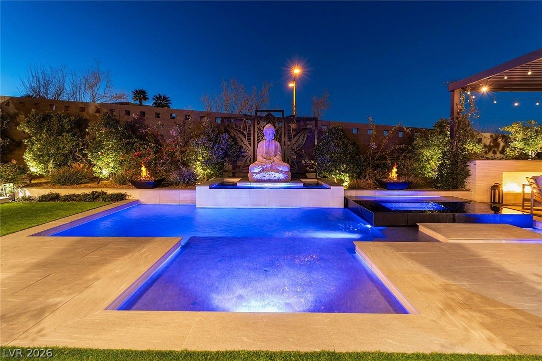 This evening shot captures a luxurious backyard oasis featuring a multi-level swimming pool and integrated spa. A central water feature showcases a serene Buddha statue set against a decorative metal screen, flanked by two fire bowls that provide a warm glow. The scene is framed by manicured landscaping and soft exterior lighting, creating a tranquil and sophisticated atmosphere.