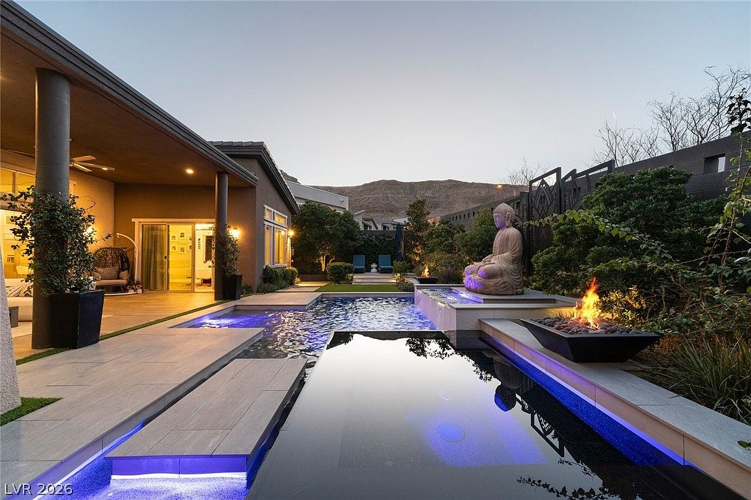 This luxurious backyard features a modern, geometric swimming pool with integrated blue LED lighting and a raised platform showcasing a large Buddha statue. A sleek fire feature adds warmth to the poolside area, while a covered patio provides a shaded transition from the home's interior. The scene is set against a desert mountain backdrop, creating a serene and sophisticated outdoor living space.