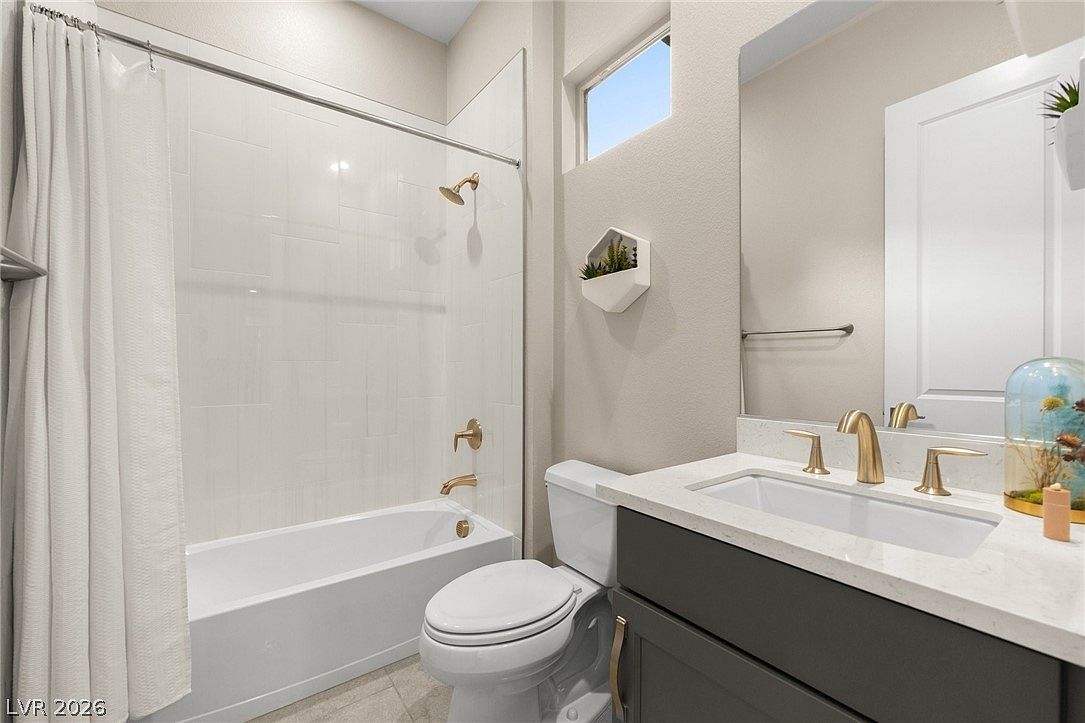 This modern bathroom features a dark gray vanity with a light-colored stone countertop and gold-toned fixtures. A white tiled shower-tub combination is positioned to the left, complemented by a white toilet and a large wall-mounted mirror. A small decorative wall planter adds a touch of greenery, while a high, rectangular window provides natural light to the space.