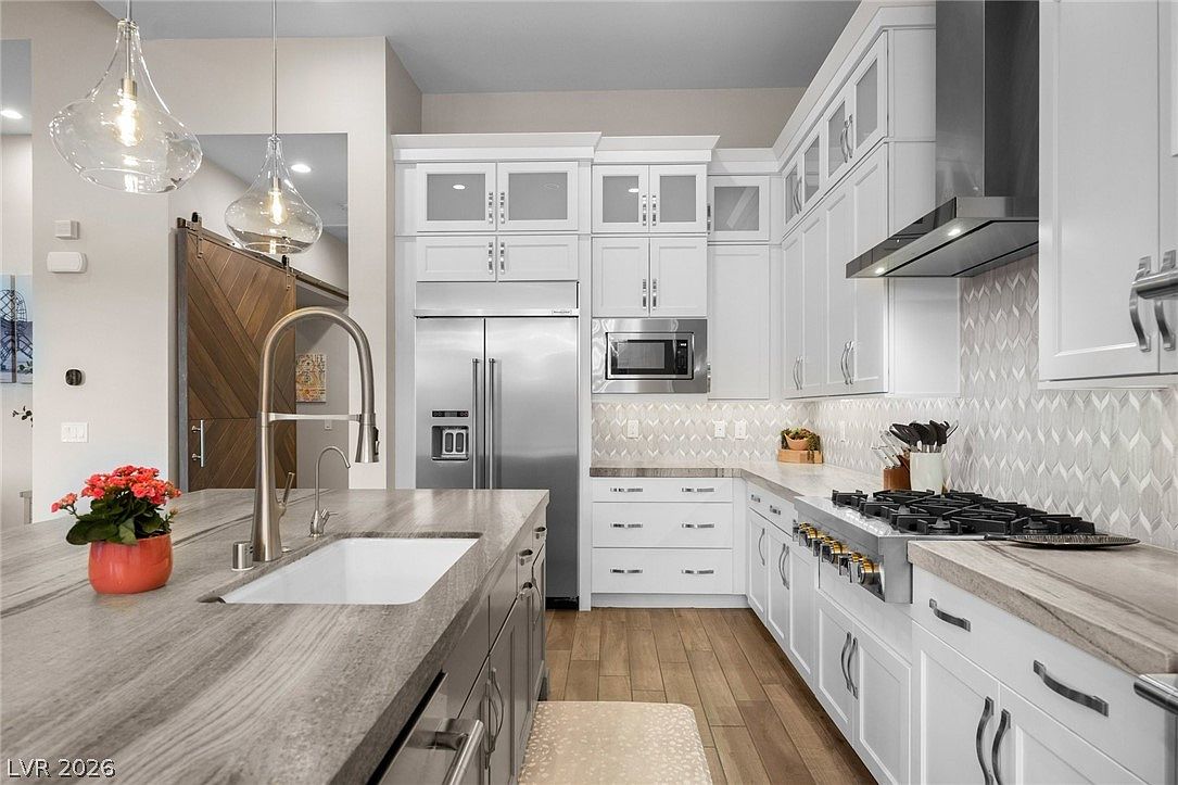 This modern kitchen features crisp white cabinetry paired with a large central island topped with a grey-toned stone surface. The space is equipped with stainless steel appliances, including a professional-style gas range and a built-in microwave, complemented by a patterned tile backsplash. Warm wood-toned flooring runs throughout the area, while two glass pendant lights hang above the island, creating a bright and inviting atmosphere.