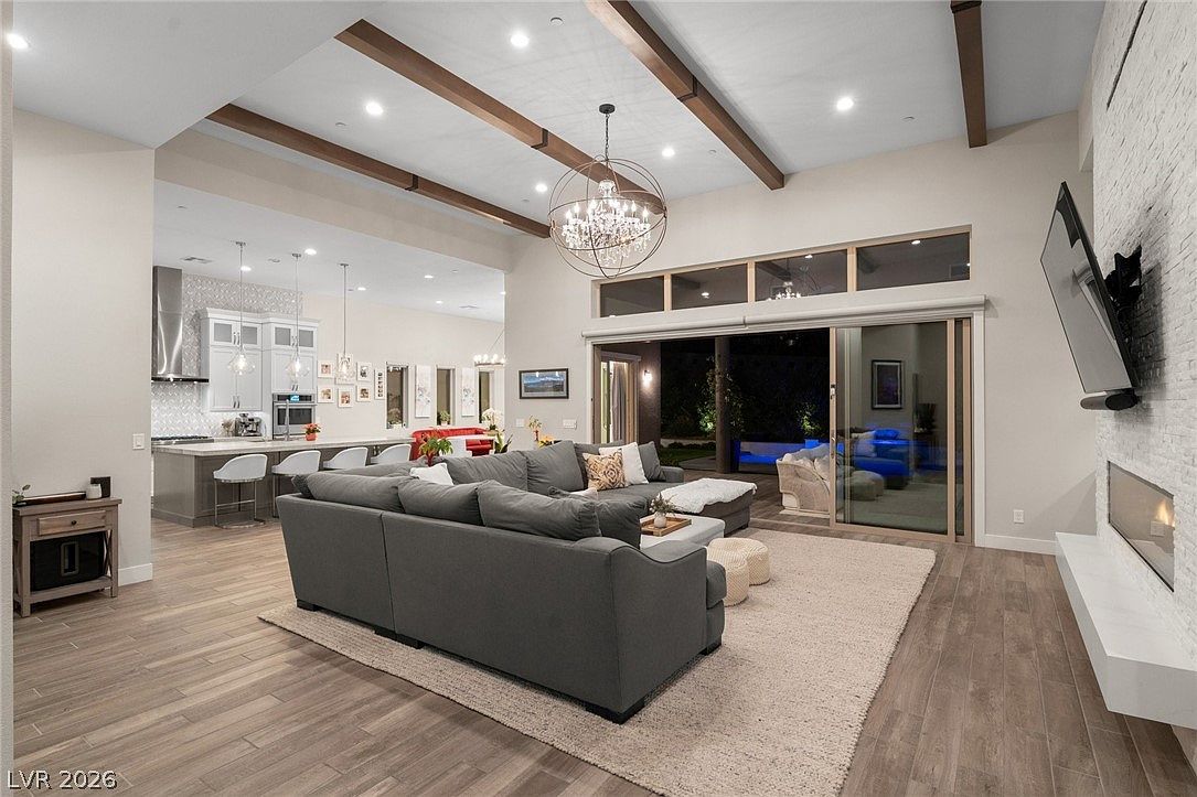 This open-concept living area features a large, plush gray sectional sofa centered on a textured area rug, facing a stone-clad chimney breast with a linear fireplace. The space is illuminated by a prominent crystal chandelier and recessed lighting, with exposed wood beams adding architectural interest to the high ceiling. Large sliding glass doors open to an outdoor patio, creating a seamless transition between the indoor living space and the exterior pool area.
