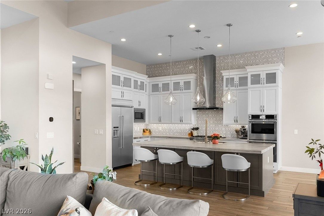 This bright, open-concept kitchen features white cabinetry, a large central island with bar seating, and stainless steel appliances. The space is accented by a patterned tile backsplash and modern pendant lighting, creating a clean and contemporary aesthetic. The view is captured from the adjacent living area, showcasing the seamless flow between the two spaces.