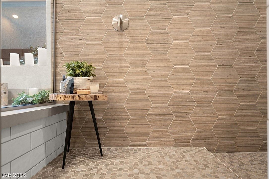 This bathroom features a striking accent wall covered in large, light-toned hexagonal tiles with a subtle wood-grain texture. A small, rustic live-edge wooden side table holds a potted plant and decorative items, positioned next to a bathtub area with a light-colored subway tile surround. The perspective is eye-level, highlighting the contrast between the geometric wall pattern and the clean lines of the adjacent fixtures.