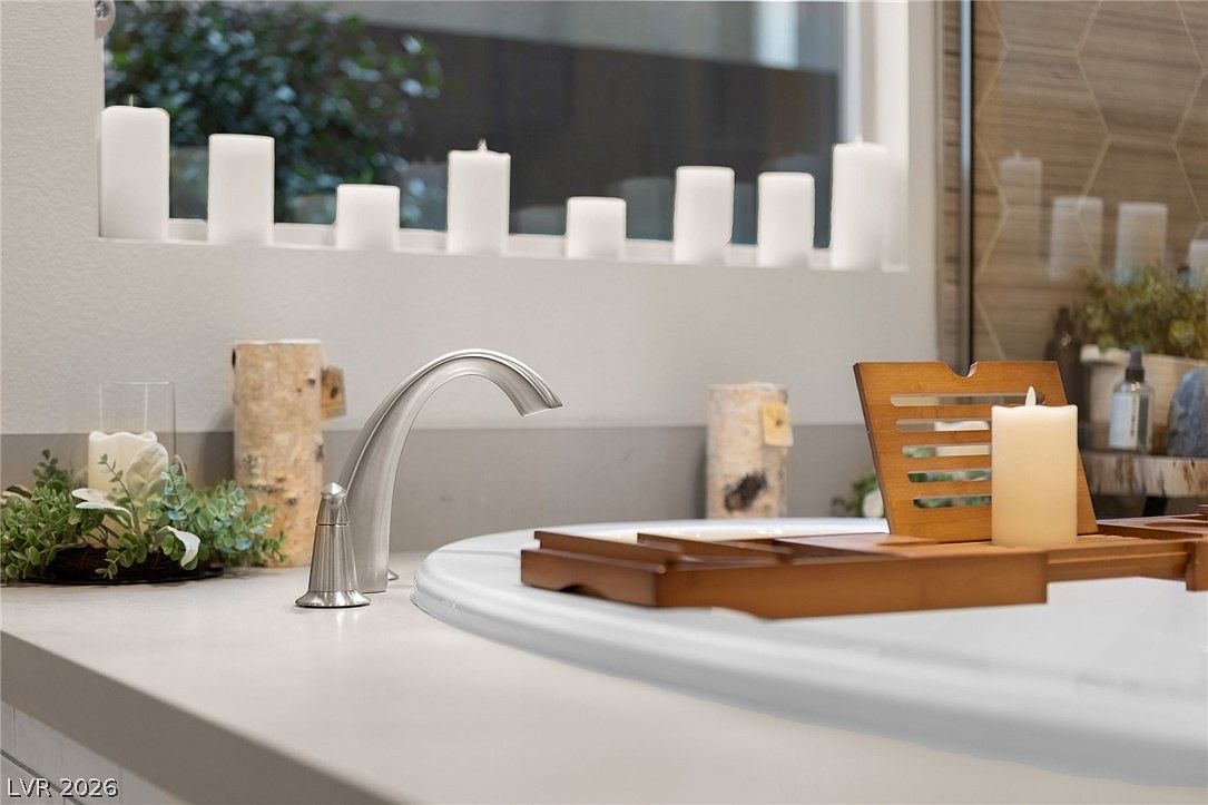 This close-up perspective focuses on a soaking tub deck, featuring a sleek, modern faucet and a wooden bath tray holding a pillar candle. The background showcases a ledge decorated with several white pillar candles and natural wood accents, creating a serene, spa-like atmosphere. The composition emphasizes a clean, tranquil aesthetic suitable for a luxury bathroom setting.