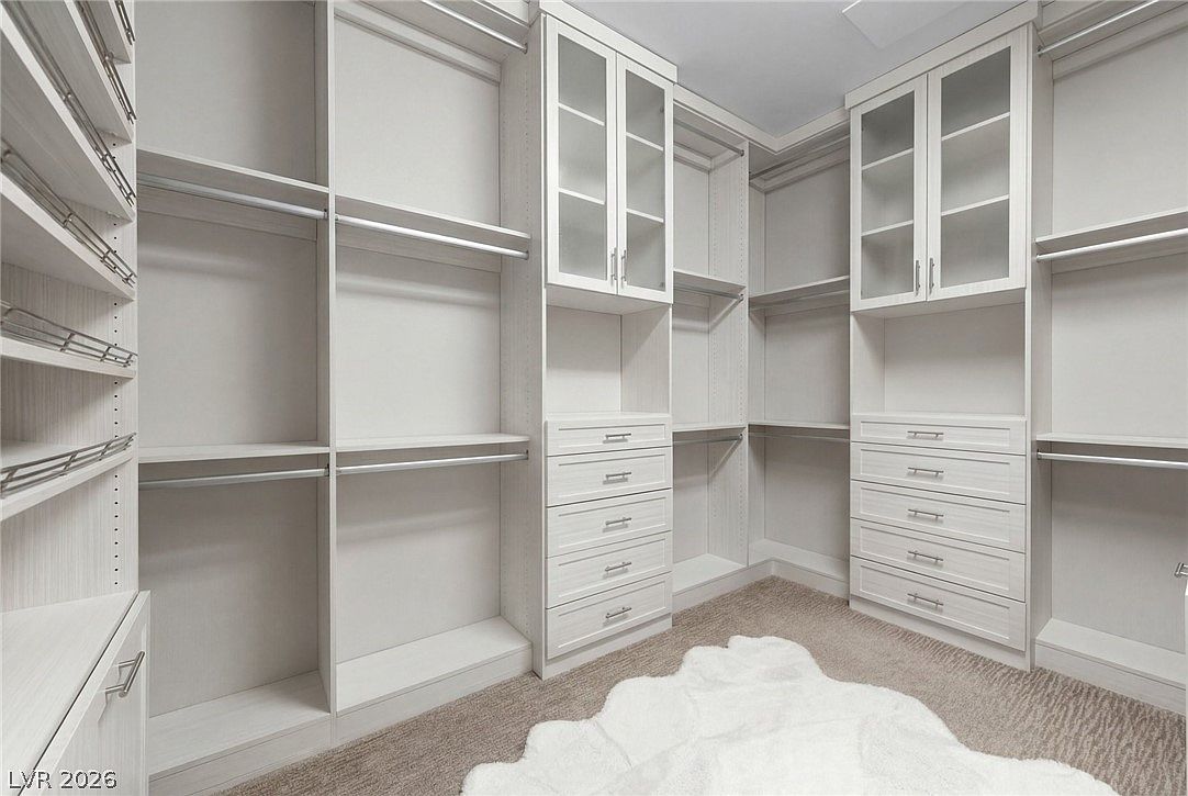 This spacious walk-in closet features custom light-toned cabinetry with a mix of open shelving, hanging rods, and glass-fronted upper cabinets. A central bank of drawers provides organized storage, while a plush, white faux-fur rug rests on the carpeted floor. The clean, neutral aesthetic creates a bright and organized atmosphere suitable for a primary suite.