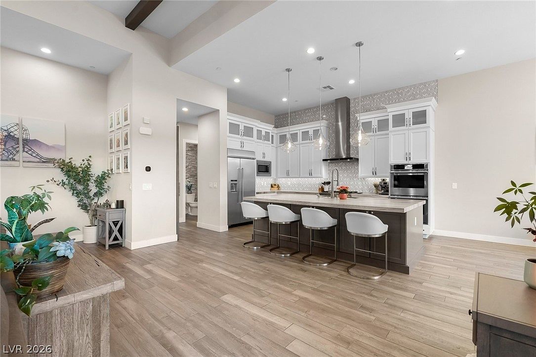 This spacious, open-concept kitchen features a large central island with bar seating and a light-toned countertop, complemented by white cabinetry and stainless steel appliances. The room is illuminated by modern pendant lights and recessed lighting, creating a bright and airy atmosphere. Wood-look flooring flows throughout the space, which transitions seamlessly into an adjacent living area, offering a clean and contemporary aesthetic.