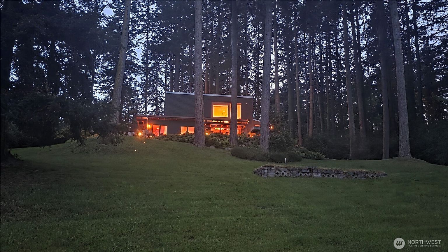 This twilight shot captures a modern, multi-level home nestled within a dense, mature forest. The structure features clean lines, dark siding, and large, illuminated windows that cast a warm glow against the darkening woods. A gently sloping lawn leads up to the house, which is framed by tall evergreen trees, creating a serene and secluded atmosphere.