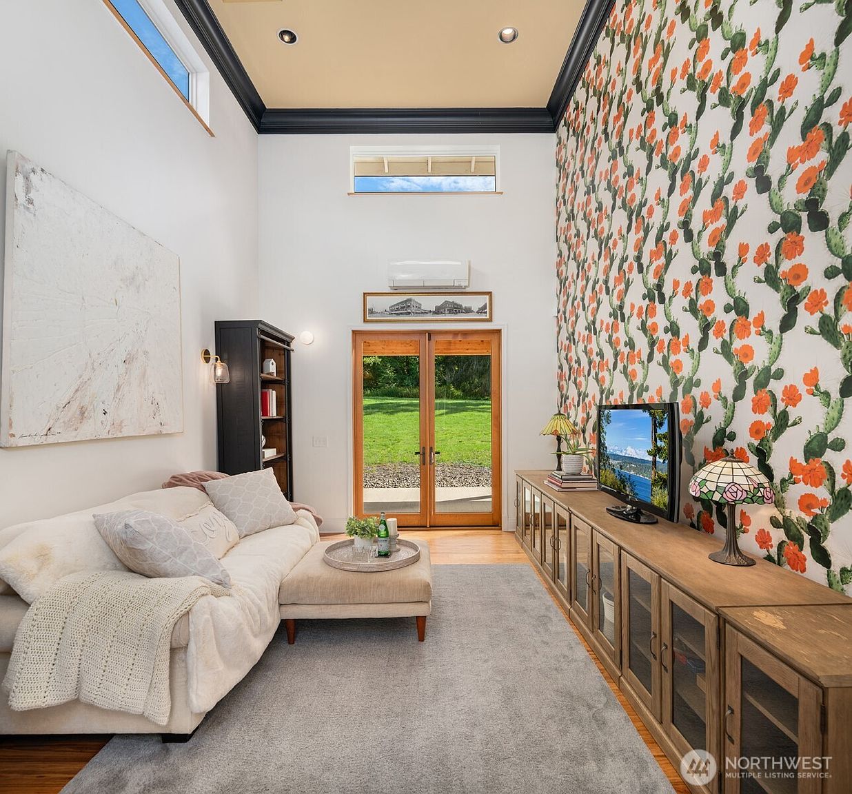 This bright living room features high ceilings with a dark trim accent and a striking accent wall covered in vibrant cactus-patterned wallpaper. The space is furnished with a comfortable cream-colored sectional sofa, a matching ottoman, and a long wooden media console topped with a television and decorative lamps. A set of French doors provides a clear view of the lush green yard, while a large abstract painting adds a modern artistic touch to the adjacent wall.