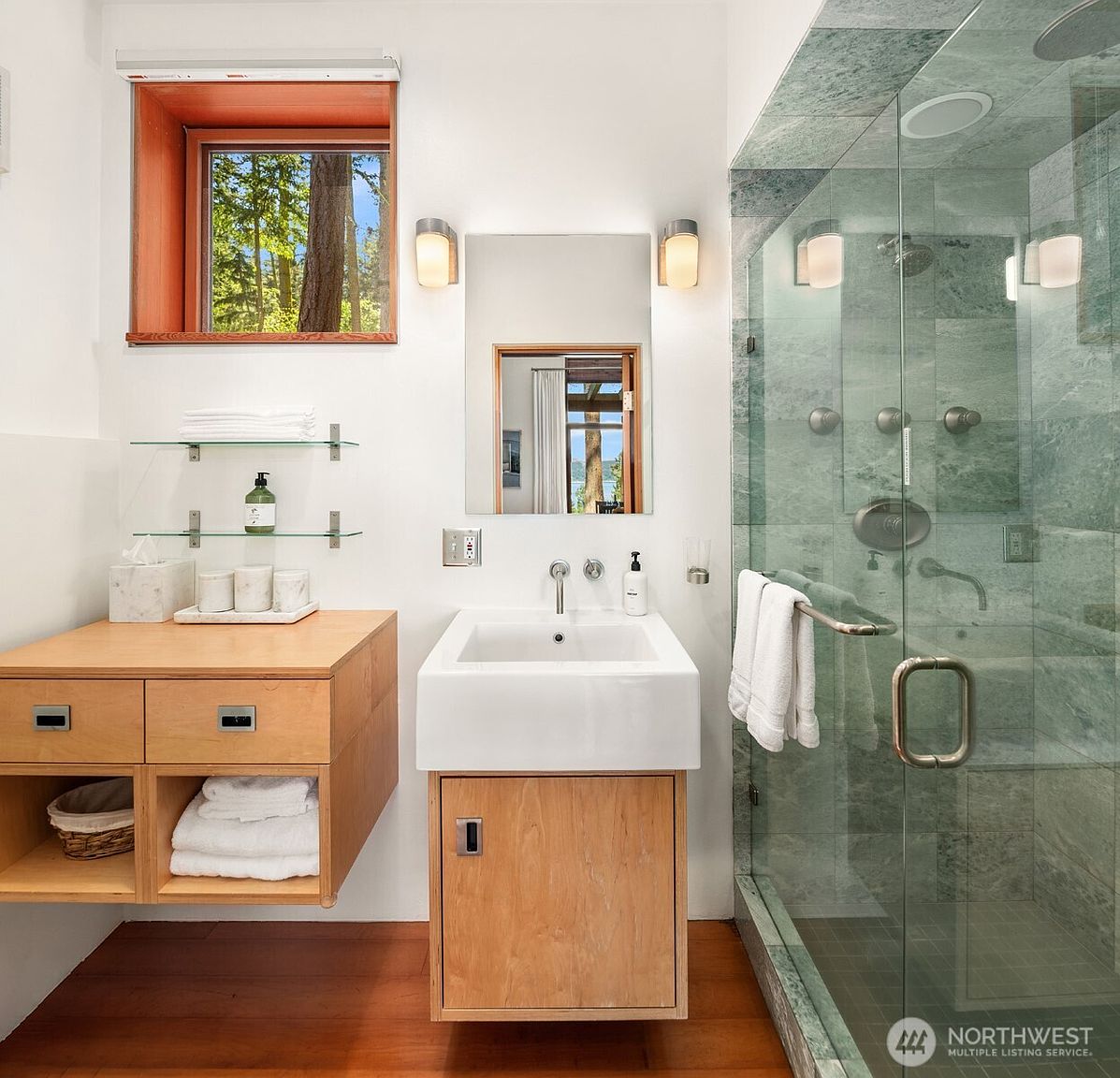 This modern bathroom features a floating light wood vanity with a white rectangular sink and a matching side storage unit with open shelving. A glass-enclosed shower with green stone-look tile walls stands to the right, while a window above the vanity provides natural light and a view of the outdoors. The space is finished with warm wood flooring and clean white walls, creating a bright and contemporary aesthetic.