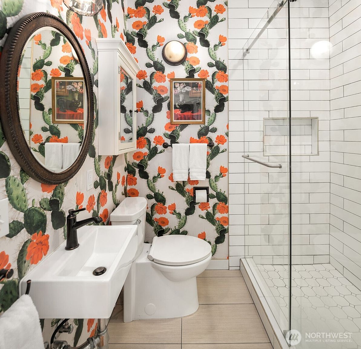 This vibrant bathroom features bold cactus-patterned wallpaper that creates a playful and eclectic aesthetic. The space includes a wall-mounted white sink with a dark faucet, a round mirror, and a walk-in shower enclosed by a glass panel with white subway tile walls. The room is finished with light-toned floor tiles and a white toilet, offering a unique and character-filled design.