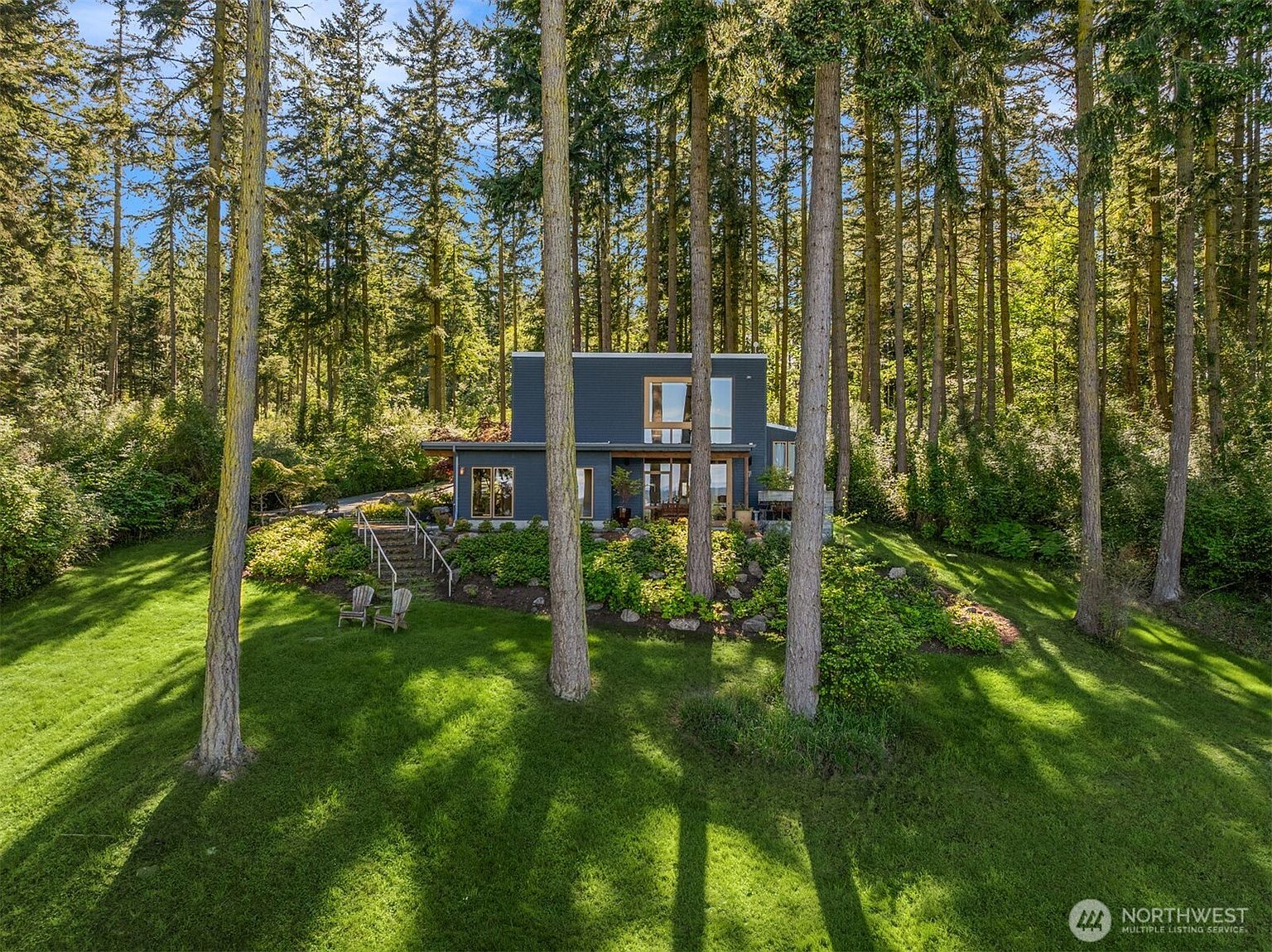 This modern, dark-sided home is nestled within a dense, lush forest, offering a serene and private retreat. The property features a tiered landscape with a grassy lawn, stone steps leading up to the house, and large windows that invite natural light into the interior. The overall impression is one of architectural harmony with the surrounding woodland environment.