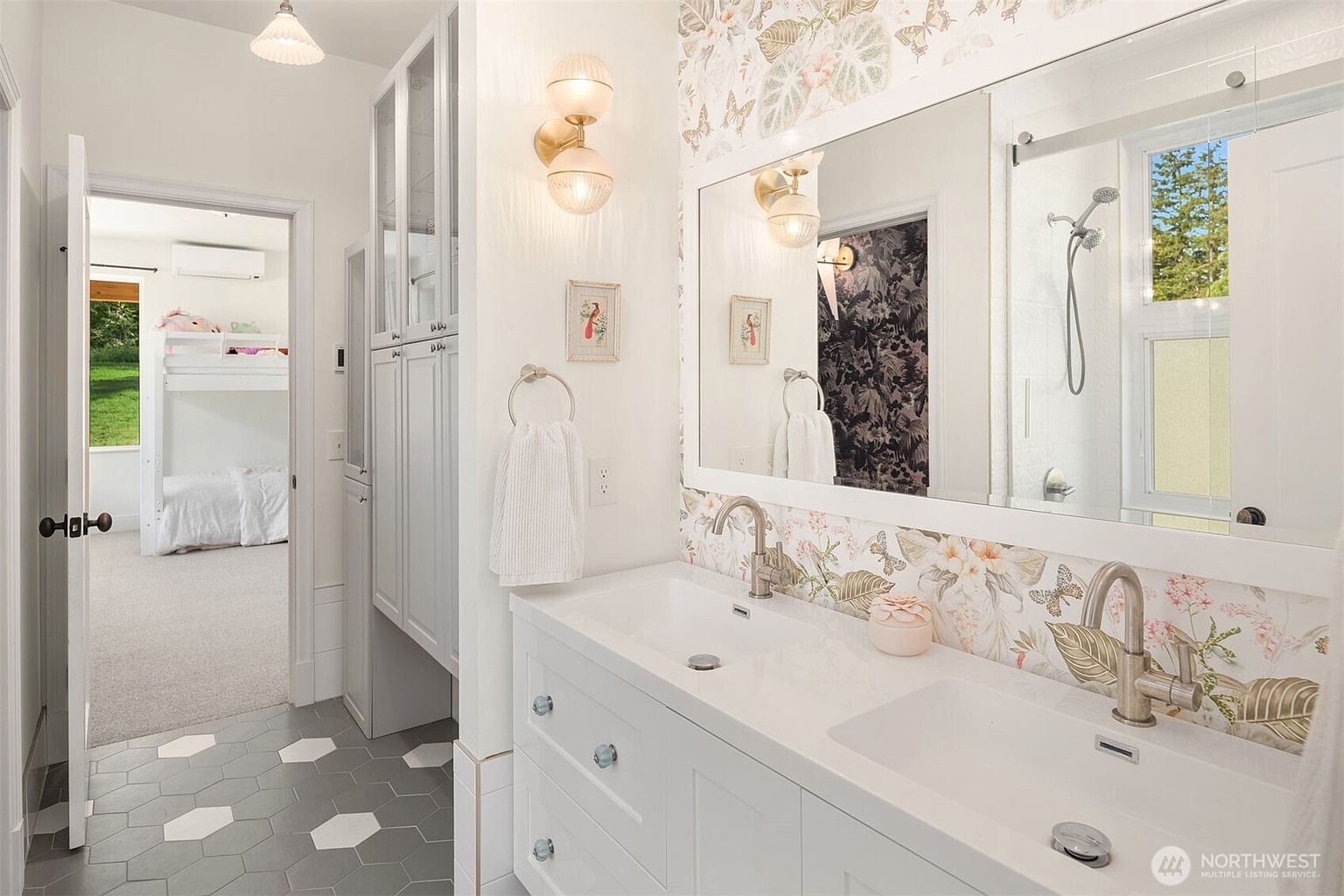 This bright and whimsical bathroom features a double vanity with white cabinetry and a patterned floral wallpaper that adds a playful touch. The space includes a glass-enclosed shower and a view through an open doorway into an adjacent bedroom with bunk beds. The flooring consists of large hexagonal tiles, and the room is illuminated by warm wall-mounted light fixtures.