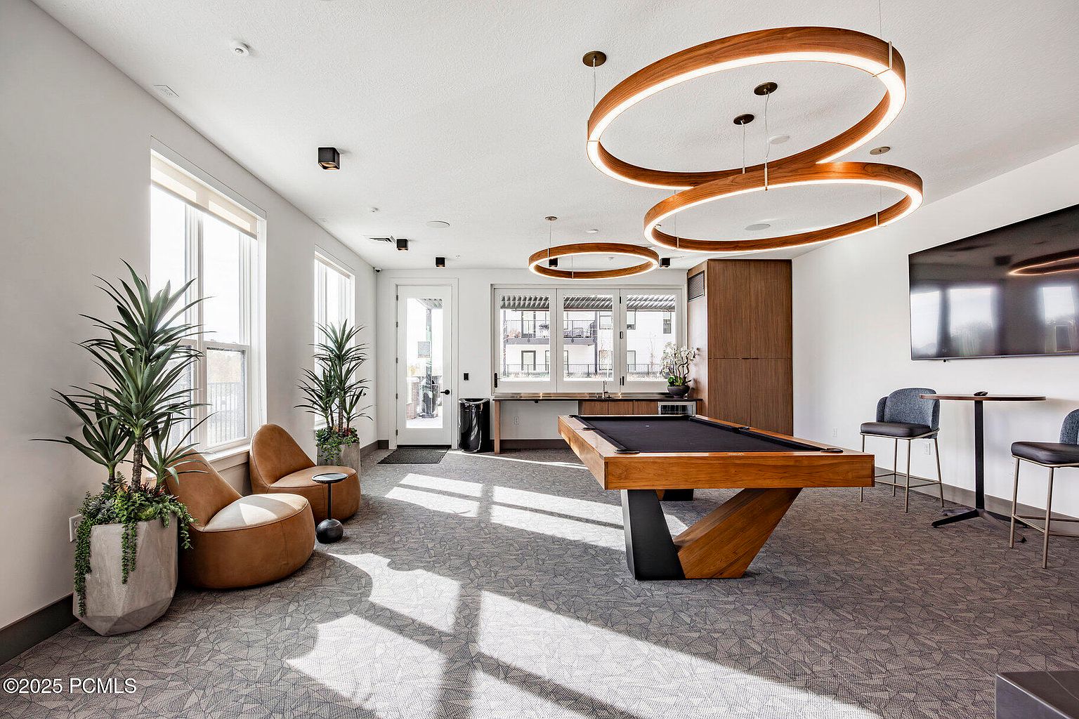 This interior shot showcases a modern amenity space, possibly a clubhouse or community center. The room features a pool table, lounge chairs, and a bar area, illuminated by unique circular pendant lights. Large windows provide natural light, and the overall design is contemporary and inviting, perfect for relaxation and entertainment.