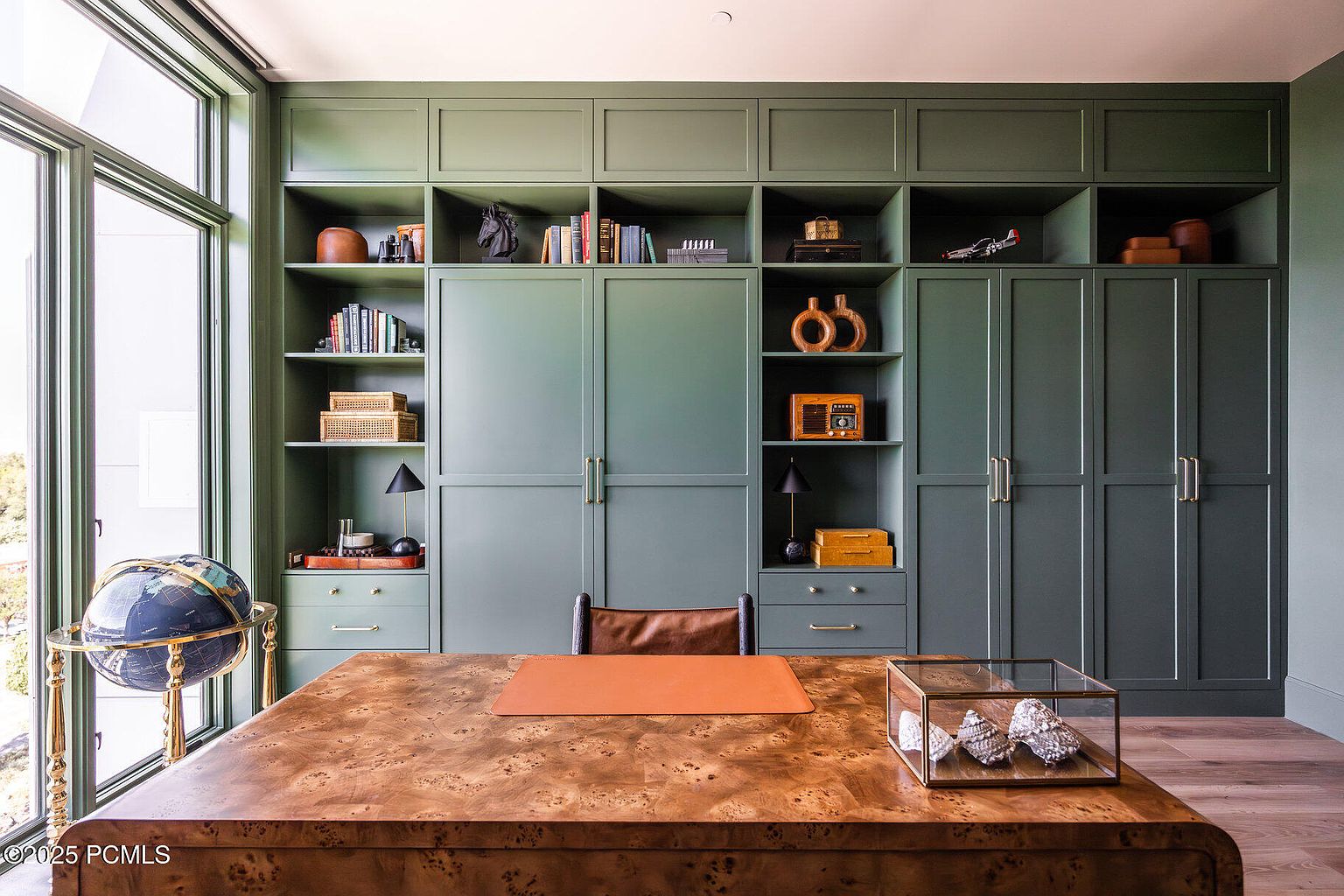 This is an interior shot of a sophisticated home office featuring custom built-in shelving and cabinetry in a muted green hue. A large wooden desk with a leather blotter and decorative objects sits in the foreground, complemented by a globe and a chair. The room is well-lit by natural light streaming through a large window, creating a warm and inviting atmosphere.