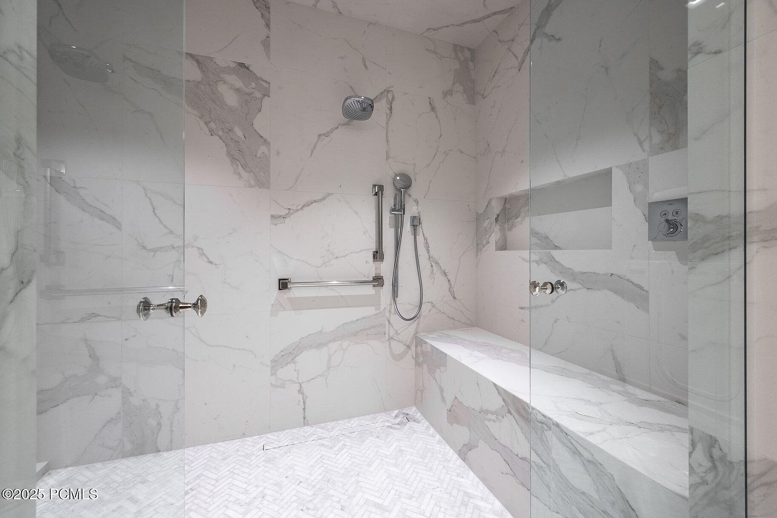 This is a luxurious primary bathroom featuring a spacious walk-in shower with marble tiling. The shower includes a built-in bench, multiple shower heads, and glass enclosure. The overall impression is clean, modern, and upscale, highlighting the high-end finishes and fixtures.